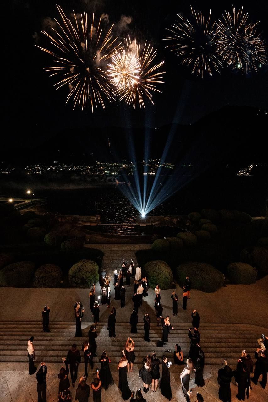 Fireworks above guests gathered on garden steps overlooking a lake at night.