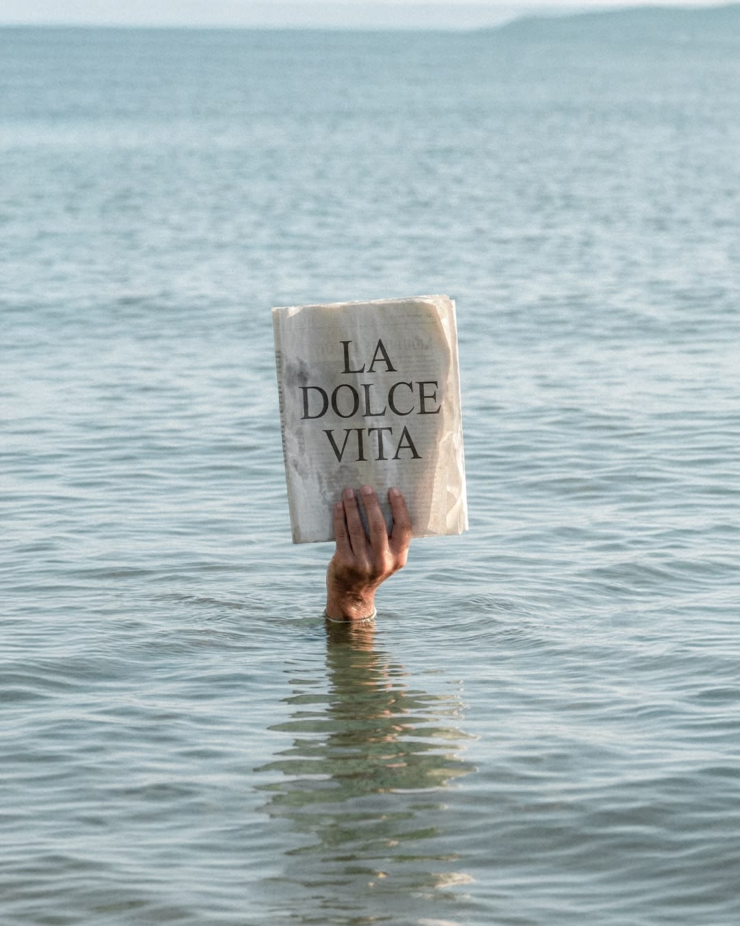 A hand holding a newspaper reading La Dolce Vita above a calm sea.
