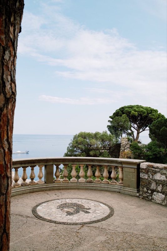 Terrasse de pierre à mosaïque de galets, pins parasols et mer calme en contrebas.
