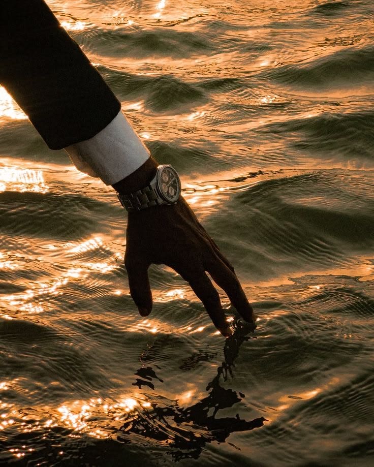 A chronograph on the wrist of a hand skimming golden water at sunset.